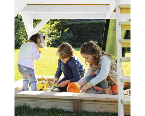 Drei Kinder spielen in einem Sandkasten mit Dach auf einer Grünfläche.