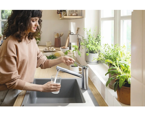 Une femme remplit un verre d''eau au niveau de l''évier avec un robinet dans une cuisine lumineuse.