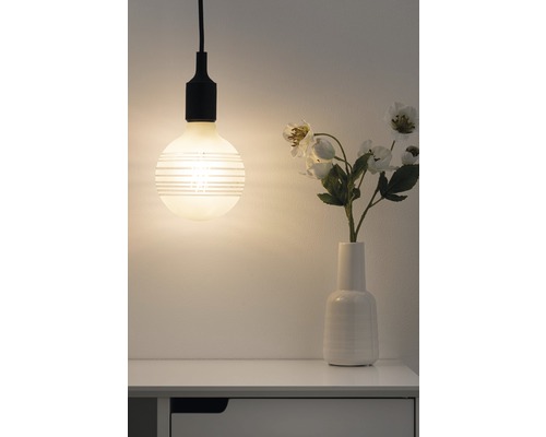 Pendelleuchte mit horizontal gestreifter Glühbirne neben einer Blumenvase auf einem Nachttisch