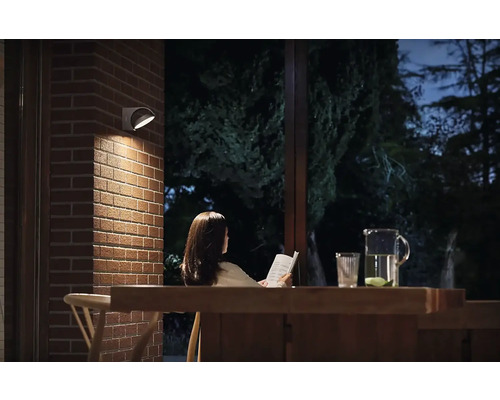 Szene mit einer Frau, die unter einer Wandleuchte im Freien ein Buch liest, auf einem Tisch stehen ein Krug und ein Glas