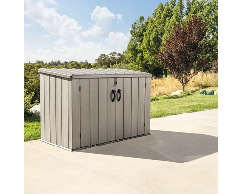 Cache-poubelle Lifetime avec ressort pneumatique 191 x 109 x 131 cm marron Coffre de jardin gris sur une terrasse avec des arbres et une pelouse en arrière-plan.