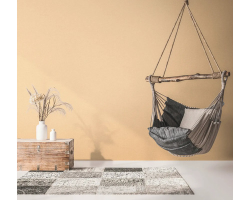 Fauteuil suspendu avec coussins, tapis à motifs et coffre en bois avec vases dans une pièce lumineuse.