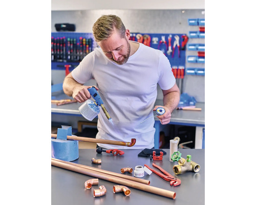 Un homme soude des tuyaux de cuivre avec un chalumeau à gaz, de la soudure, des tuyaux et des raccords sur un établi.