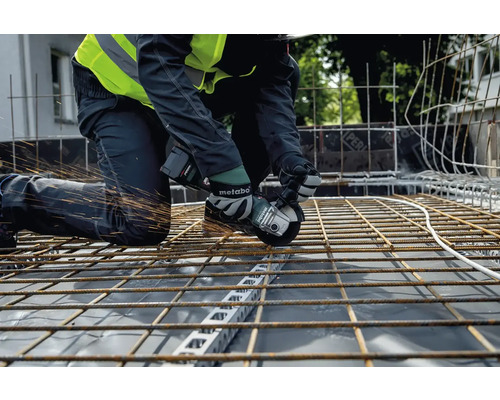 Ein Handwerker kniet auf einer Baustelle und schneidet mit einem Metabo Winkelschleifer Stahlträger, wobei Funken sprühen.