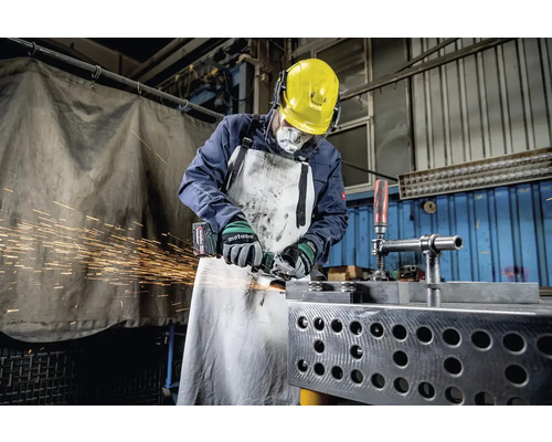 Ein Arbeiter mit Schutzhelm, Gehörschutz, Atemschutzmaske und Schürze schleift Metall mit einem Winkelschleifer, wobei Funken sprühen.