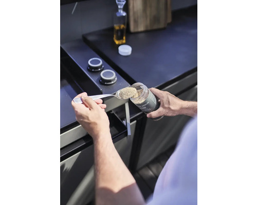 Eine Person füllt Gewürze mit einem Löffel aus einem Gewürzbehälter.