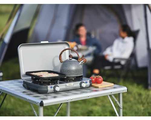 Scène de camping avec réchaud à gaz, théière et personnes devant une tente