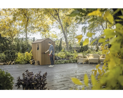 Ein Mann öffnet eine Gerätehütte im Garten, umgeben von Bäumen und Gartenmöbeln.