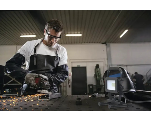 Un homme meule du métal avec une meuleuse d''angle dans un atelier, à côté de lui une lampe de travail