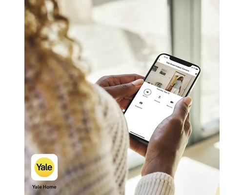 Eine Person betrachtet die Yale Home App auf einem Smartphone, das Live-Bilder einer Yale Smart Indoor Kamera anzeigt. Yale Logo.