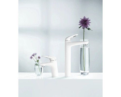 Zwei weisse Waschtischarmaturen unterschiedlicher Höhe neben Blumengestecken in Vasen