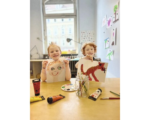 Zwei Kinder präsentieren stolz ihre mit Farbe gemalten Bilder an einem Tisch mit Farbtuben und Pinseln.