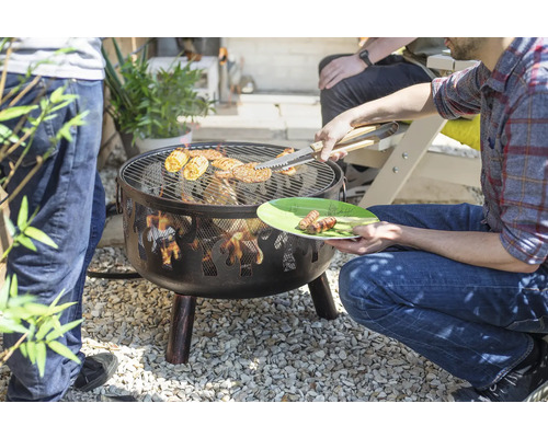 Feuerschale mit Grillrost im Garten, darauf Grillgut und eine Person mit Grillzange