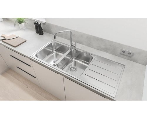 Évier en acier inoxydable avec deux bacs, égouttoir et robinet dans une cuisine claire.