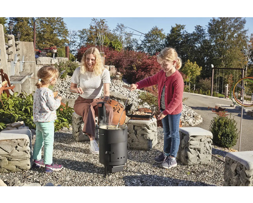 Eine Frau und zwei Kinder bereiten Stockbrot über einem Holzkohlegrill in einem Garten zu.