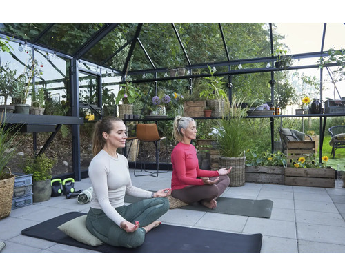 Zwei Frauen meditieren in einem Gewächshaus auf Yogamatten, umgeben von Pflanzen und Gartenmöbeln.