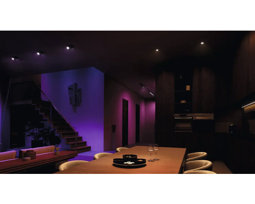 Intérieur avec escalier, table à manger, chaises et cuisine, éclairé par des plafonniers.