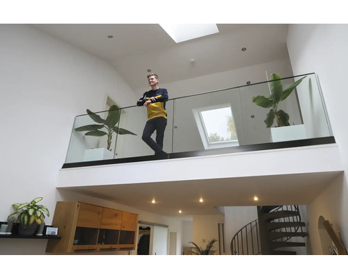 Ein heller Wohnraum mit Galerie, Glasgeländer, Zimmerpflanzen und einer Person.