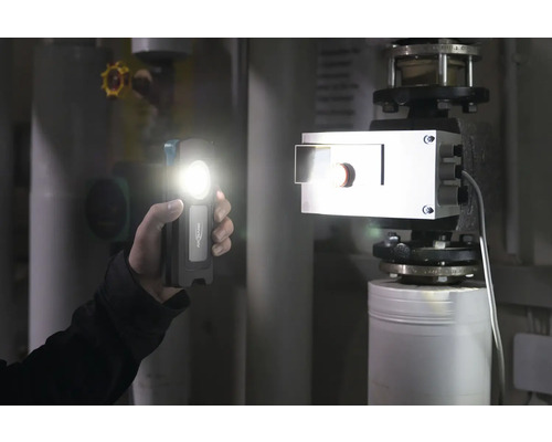 Une main tient une lampe à batterie dans un local de chauffage et éclaire un radiateur.