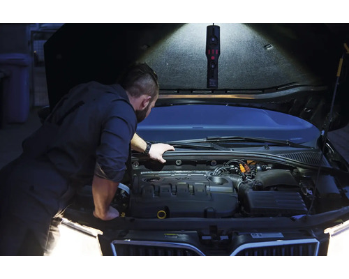 Un homme inspecte un moteur de voiture avec une lampe de travail sous le capot.