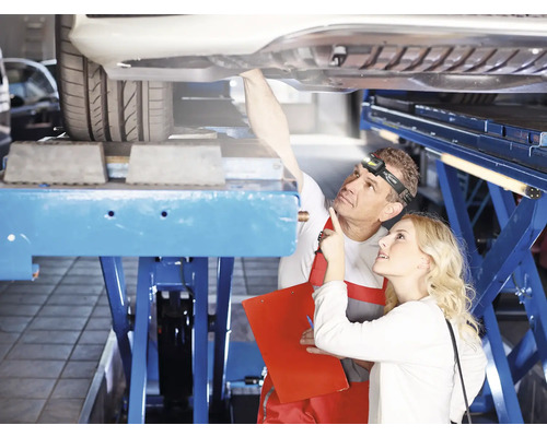 Un mécanicien avec une lampe frontale montre quelque chose à une cliente sous une voiture sur un pont élévateur.