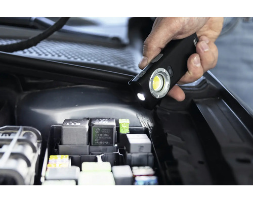 Une main tient une lampe de travail à LED au-dessus de la boîte à fusibles dans le compartiment moteur d''une voiture
