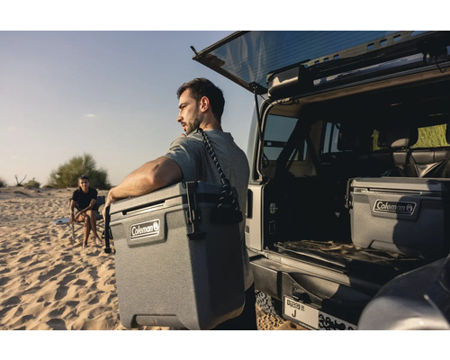 Mann trägt Coleman Kühlbox am Strand neben einem offenen Geländewagen. Eine weitere Kühlbox steht im Kofferraum.