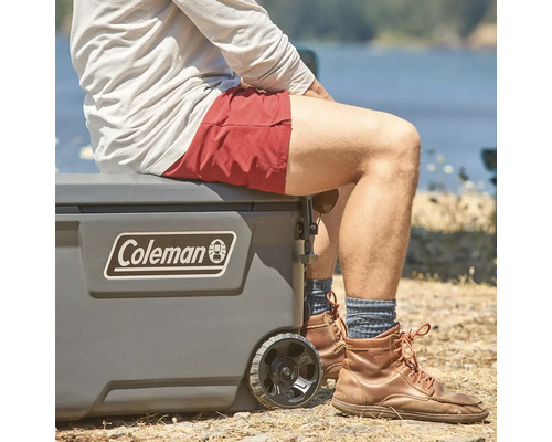Person sitzt auf Kühlbox mit Rädern und Coleman Logo.