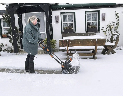 Une femme utilise une fraise à neige devant une maison en hiver.