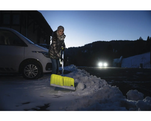 Frau schippt Schnee mit einer Schneeschaufel vor einem Auto.