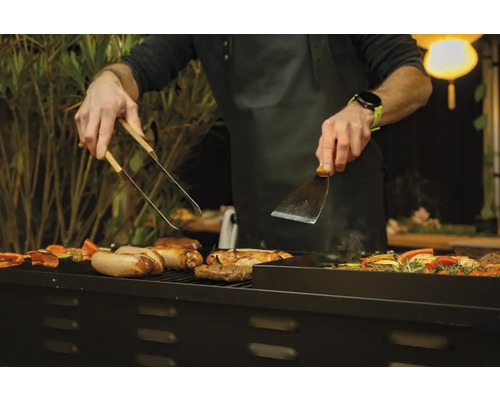 Une personne fait griller des saucisses et des légumes avec une pince à barbecue et une spatule.