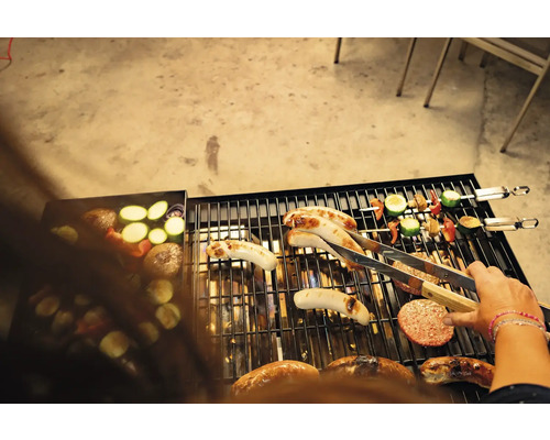 Barbecue avec saucisses, brochettes de légumes, légumes et galettes de hamburger, plus une personne tenant des pinces à barbecue.