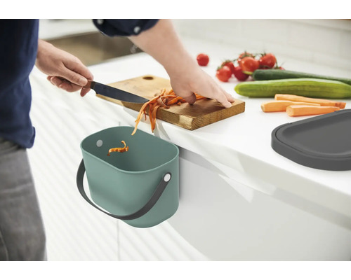 Une personne coupe des légumes sur une planche à découper en bois au-dessus d''un bac à compost dans la cuisine.