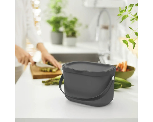 Abfalleimer mit Deckel auf einer Küchentheke neben Lebensmitteln und einer Person, die Gemüse schneidet.