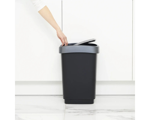 Une personne ouvre une poubelle à pédale noire avec un couvercle gris.