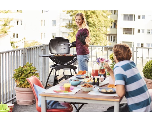 Frau grilliert mit einem Weber Grill auf einem Balkon. Es gibt einen gedeckten Tisch mit Essen und Getränken.