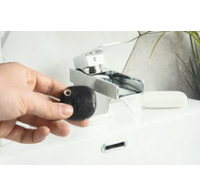 Hand hält einen schwarzen, quadratischen Wassermelder vor einem Waschbecken mit Wasserhahn