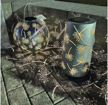 Zwei Solarleuchten aus Metall mit Löwenzahn-Muster auf Pflastersteinen.