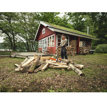 Homme avec une tronçonneuse coupant du bois devant une maison en bois avec un toit végétalisé.
