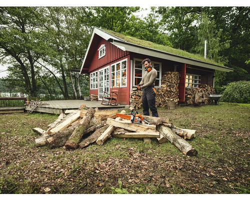 Homme avec une tronçonneuse coupant du bois devant une maison en bois avec un toit végétalisé.