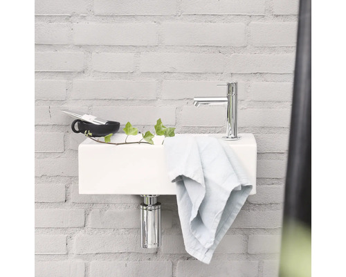 Auf einem hellen Backsteinhintergrund befindet sich ein Waschbecken mit Armatur, Handtuch, Becher und Pflanze.