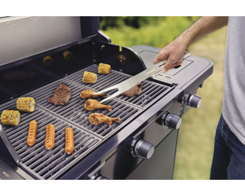 Un grill avec des aliments grillés comme des steaks, des saucisses, du poulet et des épis de maïs est retourné avec une pince à gril.