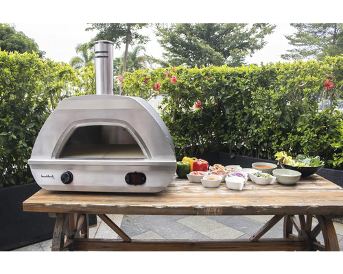 Edelstahl Pizzaofen mit Zutaten auf Holztisch im Freien.