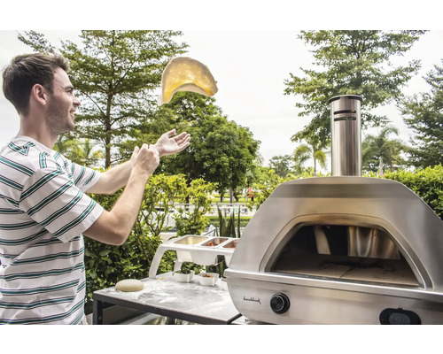 Ein Mann wirft Teig vor einem Pizzaofen im Freien in die Luft.