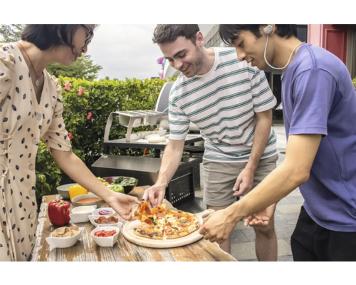 Drei Personen essen Pizza im Freien, mit einem Grill und verschiedenen Zutaten auf einem Holztisch.