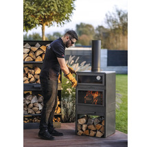 Mann mit Schutzhandschuhen bereitet einen Cook King Grillofen im Garten vor. Holzstapel neben dem Ofen.