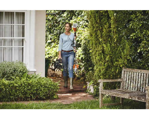 Une femme porte une débroussailleuse à batterie et un taille-haie à batterie dans un jardin bien entretenu