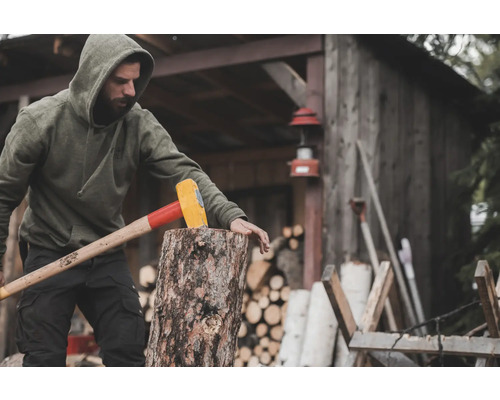 Mann spaltet Holz mit einem Spalthammer vor einer Holzhütte.