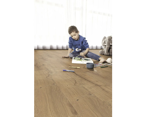 Kind malt auf einem Holzfußboden. Im Hintergrund befindet sich ein Plüschtier-Elefant.