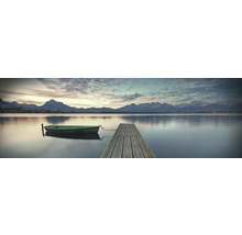 Ein ruhiger See mit einem Holzsteg und einem kleinen Boot vor einer Bergkulisse.